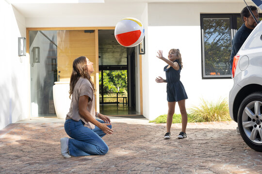 African American family kneeling, reaching, tossing colorful beach ball by white car on driveway