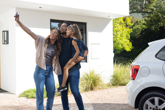 Diverse family with child taking selfie with phone on driveway by white car and modern house