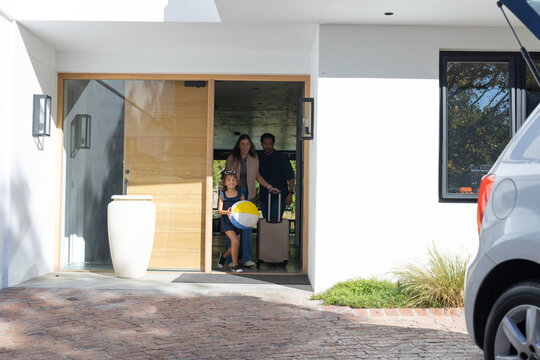 African American family leaving front door, child holding beach ball, dad pulling suitcase