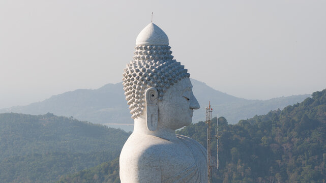 Aerial view of the majestic Big Buddha statue in profile against a backdrop of lush green hills and a hazy sky in Tambon Karon, Chang Wat Phuket, Thailand.