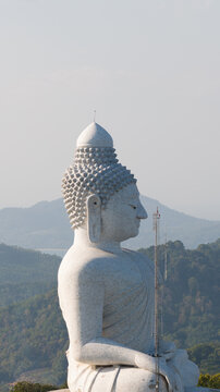 Aerial view of the white marble Big Buddha statue in profile against a backdrop of lush tropical hills and a hazy sky in Tambon Karon, Chang Wat Phuket, Thailand.