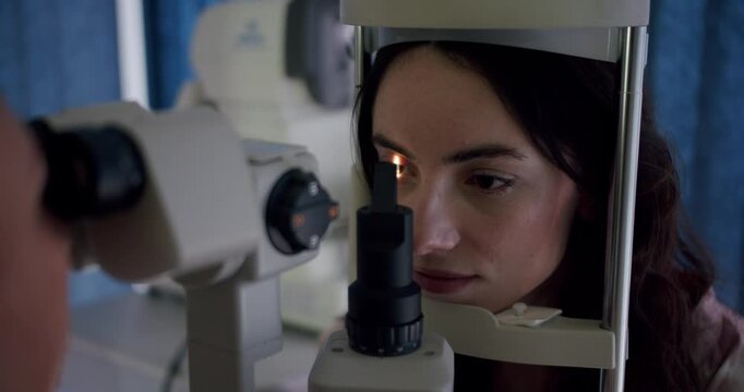 A focused young woman receiving an eye test, looking through advanced optical equipment in a clinical setting, highlighting health care and technology. Young Woman Undergoing Eye Examination in Clinic