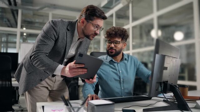 Two diversity business men reviewing information in digital tablet and computer laughing funny happy talking Caucasian man boss showing online data to Arabian intern laugh talk fun joke in office