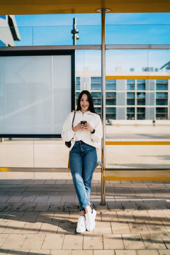 Young woman with dental braces holding smartphone and looking away, career growth, ambition, successful digital nomad waiting at city bus stop, wearing white sweater and jeans, urban lifestyle.