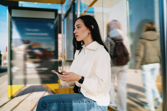 Thoughtful woman holding smartphone at city bus stop with blurred people in background, waiting for commute, looking away, urban transportation, street lifestyle, candid photography style.