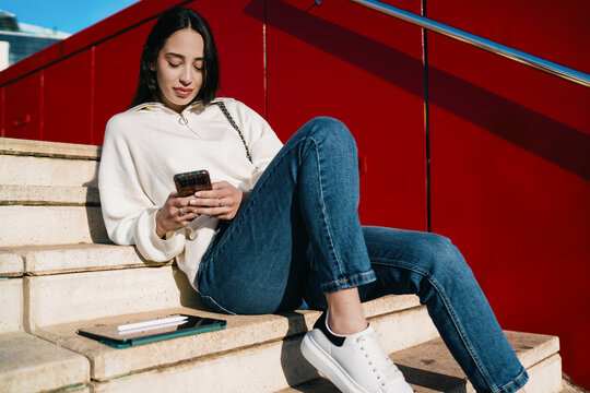 Young lady wearing earbuds and holding mobile phone while waiting for commute, listening to audio book or podcast, urban travel, serenity, mindfulness, contemporary digital lifestyle, city day.