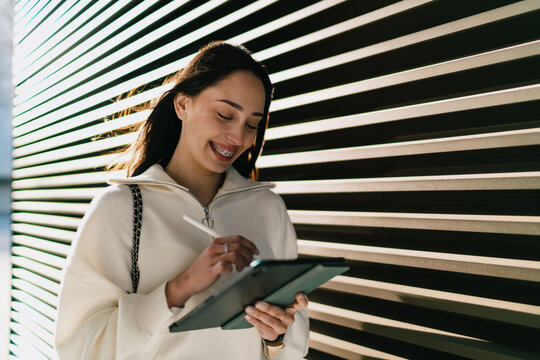 Happy young woman with braces using tablet and stylus for creative work outdoors. Smiling female student or freelancer drawing on digital device, online education and digital nomad lifestyle.