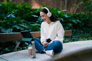 Fototapeta premium Cheerful girl with braces in wireless headphones sitting cross-legged on bench and using tablet. Happy woman enjoying audio content or video call in urban green park, digital nomad representation.