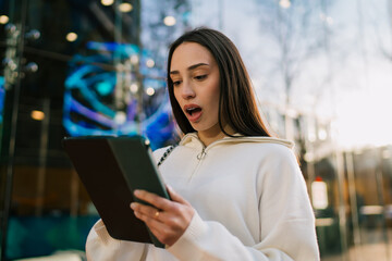 Surprised young woman with braces looking at digital tablet in urban city setting. Excited girl winning or receiving great news on mobile device outdoors, success and modern connectivity concept. © BullRun