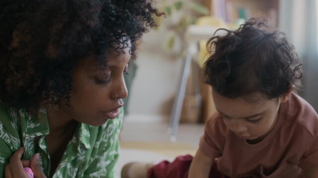 Handheld close up shot of toddler sitting next to African American mother drawing on paper with pencils while bonding at home