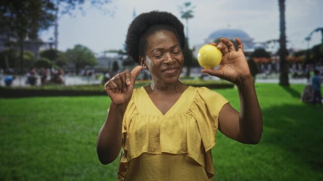 Woman holding lemon between fingers and smiling while pinching near building facade in a grassy public space; freshness joy.