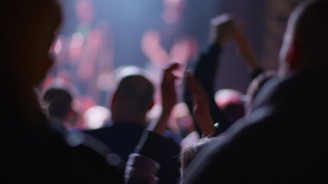 Silhouetted fan claps hands above head holding object, warm red and pink bokeh stage lights fill background, crowd visible around in dark