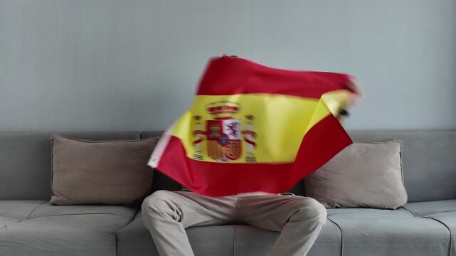 Emotional man sitting on a sofa watching a football match, waving the national flag of spain and screaming with joy for his team's victory