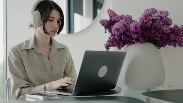 Young woman wearing headphones working on her laptop at home, with purple flowers beside her. A focused and relaxed work-from-home environment. Woman Working at Home