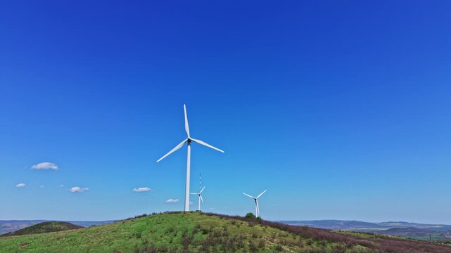 Three wind turbines rotate on a green hilltop with a bright blue sky overhead. The landscape shows rolling hills and a few distant clouds. Natural energy generation is the focus here.