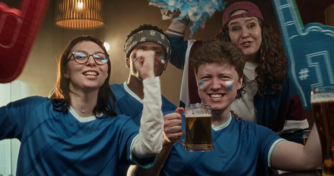 Portrait shot of four happy friends in fan makeup wildly celebrating their teams victory, posing with foam fingers, pom-poms, and beer at bar