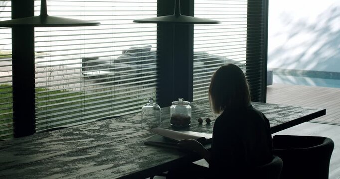 A serene morning scene of a young woman reading a book by a large window with blinds, overlooking a sunny poolside. Woman Reading Book in Modern Home Interior with Pool View
