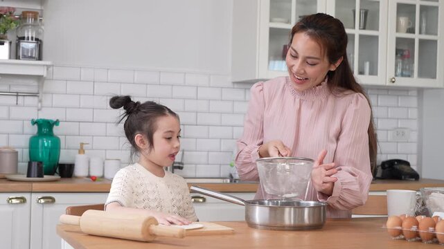 Asian happy mother and little daughter sifting flour with sieve into bowl in kitchen, Family home cooking, baking bakery, childhood learning, motherhood bonding, domestic lifestyle food concept