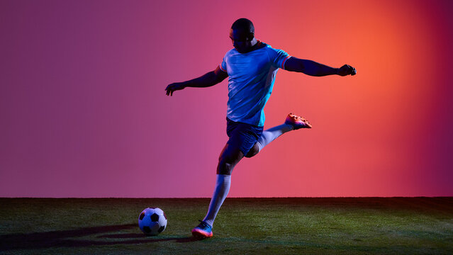 Football player preparing penalty kick on grass field with neon gradient background. Concept of sports promo, match preview, training campaign and football equipment advertising.