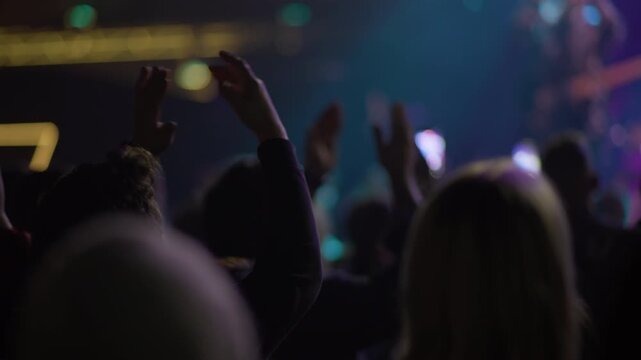 Concert fan raises both arms overhead, teal stage atmosphere visible behind, blurred neon sign strip upper left, other fan and performer visible in background