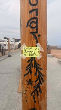 A wooden post with decorative carvings and a yellow sticky note that reads 'You look amazing & don't forget!' in a coastal area.
