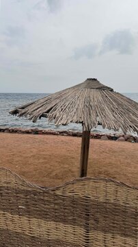A thatched umbrella stands on a sandy beach near the ocean. The sky is overcast, and gentle waves lap at the shore.