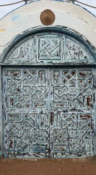 An ornate blue wooden door with intricate carvings and a circular emblem at the top. The door shows signs of weathering and age.