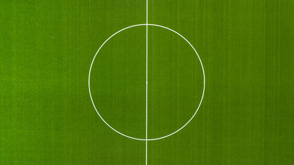 Aerial closeup of a vibrant green football pitch featuring the center circle and halfway line. The white markings contrast against the textured turf, symbolizing the start of a match. Zenithal view. © Stefano Tammaro