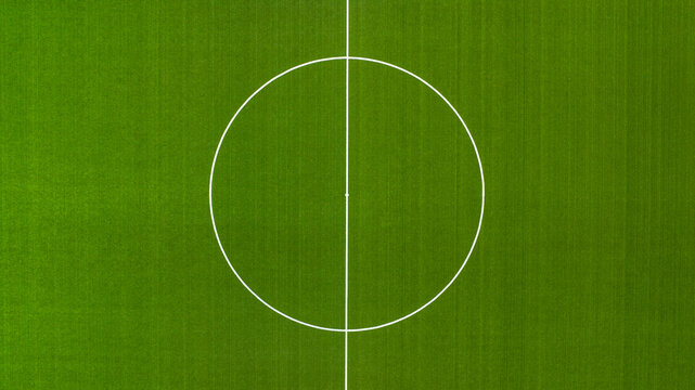 Aerial closeup of a vibrant green football pitch featuring the center circle and halfway line. The white markings contrast against the textured turf, symbolizing the start of a match. Zenithal view.