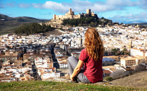 young woman sitting on a hill and contemplating the panoramic view of alcala la real and the historic fortress of la mota, spain