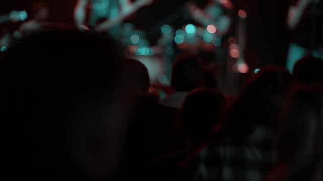 Concert fan raises and claps both hands overhead at live music event, blue stage light surrounds crowd from front, wristband visible on arm