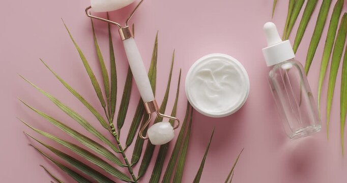 Top view White bottle, serum, facial pink roller with palm leaf rotate for cosmetics on pink background in studio. Natural cosmetics production for hair and skin care. Different cosmetic bottles.