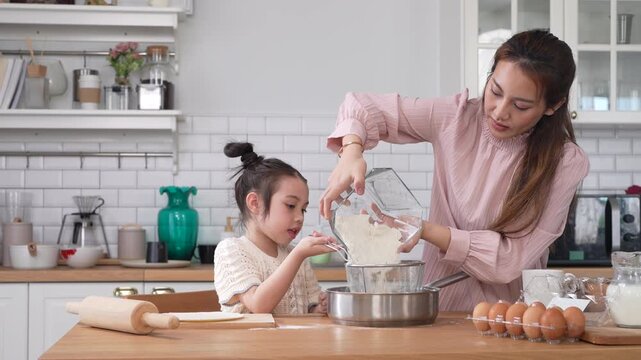 Asian happy mother and little daughter sifting flour with sieve into bowl in kitchen, Family home cooking, baking bakery, childhood learning, motherhood bonding, domestic lifestyle food concept
