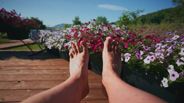 Feet rest bare on wooden deck beside vibrant flower beds. Sunlight warms skin, calming mood. Peaceful outdoor relaxation, perfect for wellness or travel content