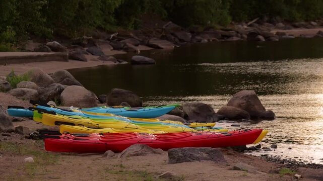 Serene lakeside scene features a lineup of colorful kayaks paused on the rocky shore. The calm water mirrors the soft glow of the setting sun, while large boulders and lush greenery frame the peaceful
