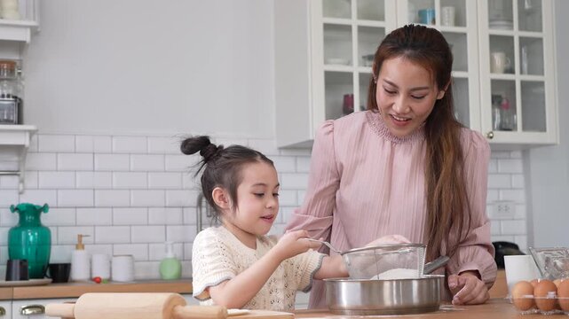 Asian happy mother and little daughter sifting flour with sieve into bowl in kitchen, Family home cooking, baking bakery, childhood learning, motherhood bonding, domestic lifestyle food concept
