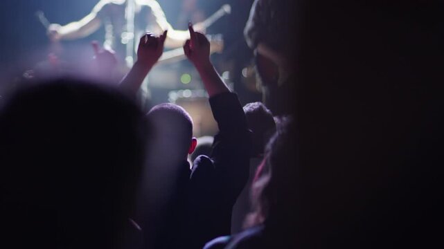 Fan with shaved head raises fist among crowd at live event, red stage light illuminates scene dramatically, blurred guitarist and bass player on stage behind