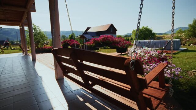 Wooden porch swing gently sways under bright sun. Flowers bloom in vibrant colors around swing. Peaceful rural view with distant house and hills. Calm, relaxing scene perfect for home, vacation