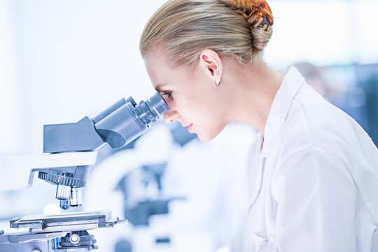 Medical pathologist looking through microscope eyepiece for cancer diagnosis. Female scientist in safety glasses analyzing biopsy slide in sterile hospital lab room.
