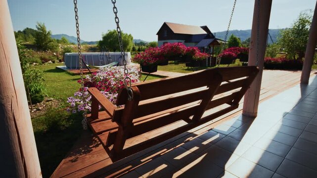 Wooden swing hangs on porch, swaying gently. Sunlight streams through, casting long shadows. Vibrant pink flowers bloom around the swing. Green lawn and distant hills create peaceful backdrop