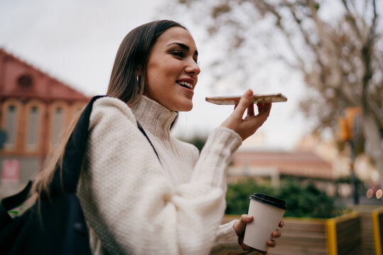 Happy female student using voice command on smartphone holding coffee to go, young woman in urban environment communicating through mobile application, digital nomad and technology concept.