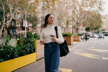 Fototapeta premium Attractive millennial girl walking city street holding smartphone and coffee cup, young woman in stylish white sweater and backpack looking away, urban youth lifestyle and digital nomad concept.