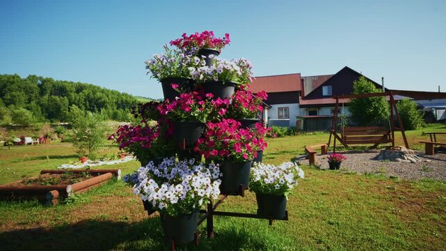 Tall tiered planter holds vibrant pink and white flowers. Sunny backyard garden with wooden swing and cottage in background. Grass lawn stretches under clear blue sky. Ideal for home decor, gardening