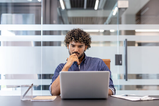 A thoughtful young Indian man sits at a desk in the office and looks thoughtfully at the laptop screen
