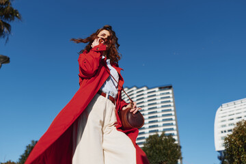 Fototapeta premium A stylish woman in a red coat walks outdoors in an urban setting, sunlight bright, wind blowing her hair. A bold fashion moment highlighting confidence, street style, and contemporary city living.