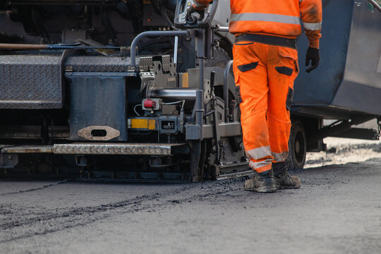 Worker operates asphalt paving machine on road construction site in city during sunny day