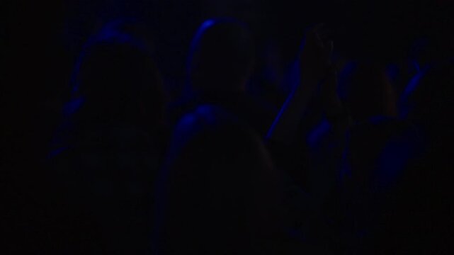 Woman silhouette clapping at live music concert under intense blue stage light in dark venue, slow motion, crowd atmosphere