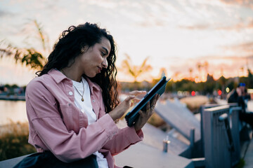 Young woman taps tablet screen with intent, focused and engaged, reflecting seamless digital fluency, mobile concentration and independent tech flow at sunset near city harbor. © BullRun