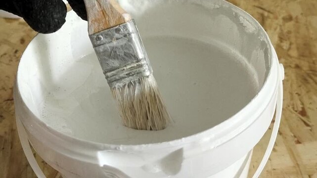 A painter's hand in a glove dips a brush in white paint in a jar. Repair, painting, whitewashing