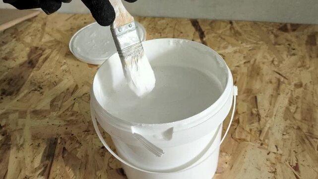 A painter's hand in a glove uses a brush to stir the white paint in the jar.